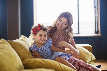 美丽的微笑的母亲与她的小可爱的女儿在蓝色礼服愉快地花费时间一起在沙发在家