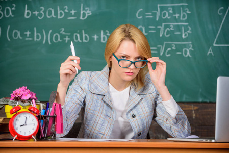 女教师坐桌教室黑板背景。老师可疑地偷看了眼镜。她对你的报告或知识有疑问。老师怀疑你的知识和技能