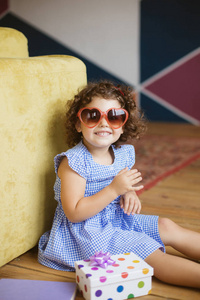 可爱的小微笑的女孩与深色卷曲的头发在蓝色礼服和太阳镜愉快地看在照相机与礼物盒在地板在家