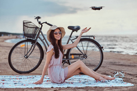 一个年轻的黑发女孩在太阳镜和帽子穿着礼服的肖像享受在海滩上度假, 玩 quadcopter