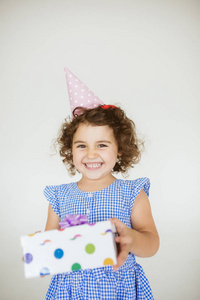 欢快的女孩与深色卷曲的头发在蓝色礼服和生日帽高兴地看着在相机与礼品盒在手上白色背景