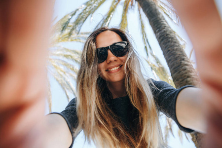 年轻美丽的女人游客或博客制作一个自拍或向其订户报告在炎热的国家棕榈树的背景下