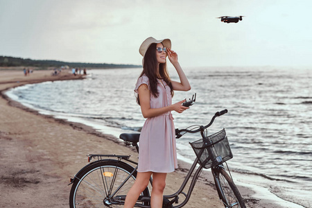 迷人的女孩在太阳镜和帽子穿着礼服享受在海滩上的假期, 玩 quadcopter