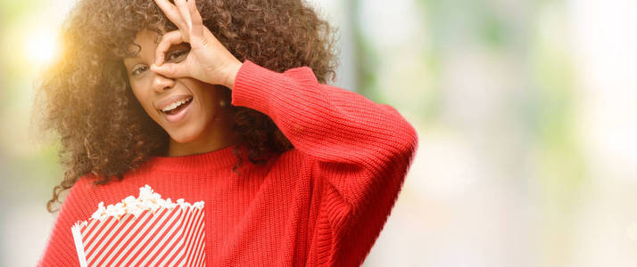 非洲裔美国妇女吃爆米花与愉快的面孔微笑做 ok 标志用手在眼睛看通过手指