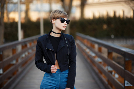 时尚的年轻女子在黑色的太阳镜黑色夹克与牛仔裤步行户外照片