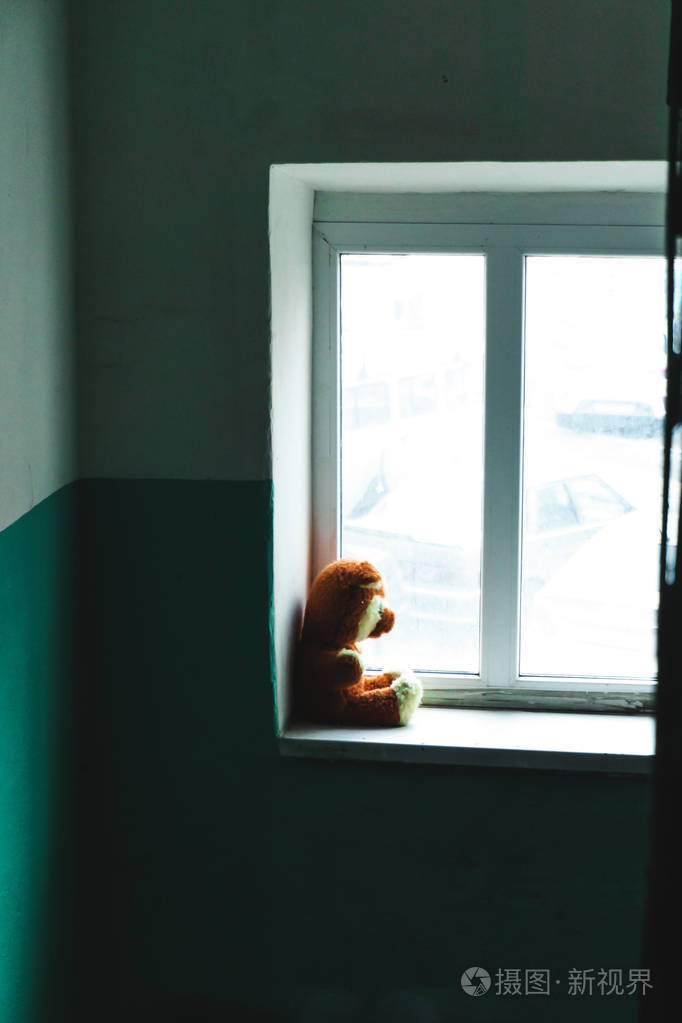 遗弃儿童玩具泰迪熊不开心的孩子坐在窗台上独自一人在门口, 看着窗外