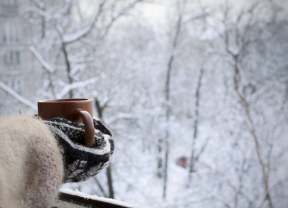 手拿着针织手套, 在被雪覆盖的树的背景上喝一杯热饮