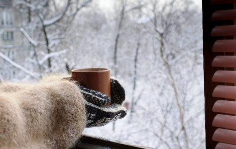 手拿着针织手套, 在被雪覆盖的树的背景上喝一杯热饮