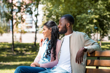 微笑的非洲裔美国人夫妇坐在木凳上公园, 看着远离