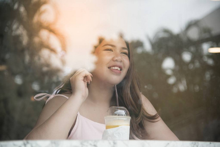 美丽的亚洲胖女人的肖像喝橙汁和使用手机在咖啡馆里