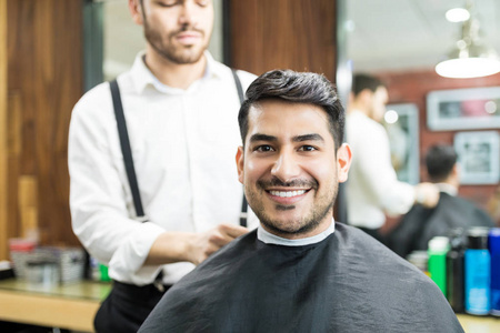 迷人的男性客户微笑, 而理发师调整美容披肩在他的沙龙