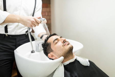 放松的年轻人, 眼睛闭上倚在水槽, 而理发师洗他的头发在沙龙