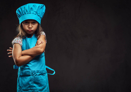 不满的小女孩身着蓝色厨师制服站在交叉的武器。在深色纹理背景下隔离