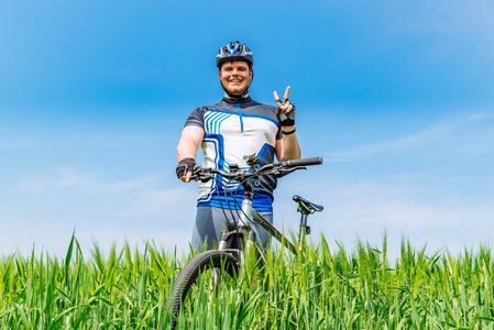年轻成人微笑的人站立与自行车在绿色大麦在背景蓝色天空申请了
