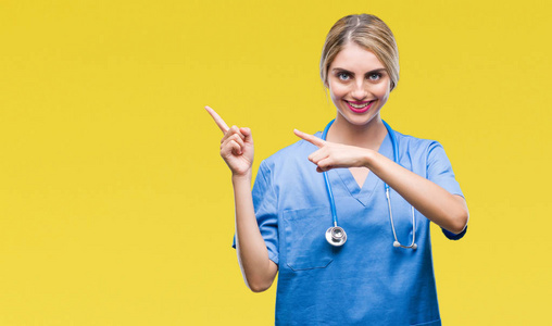年轻美丽的金发医生护士妇女在孤立的背景微笑, 看着相机指向用两只手和手指的一侧