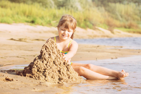 一个金发碧眼的小女孩穿着泳装, 在夏天阳光明媚的天气里在河岸上建造一座沙子城堡。