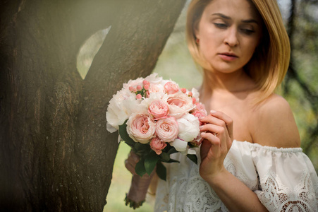 一个年轻的金发女人在白色的礼服和一束鲜花站在树上的招标肖像
