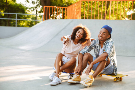 一个快乐的年轻非洲夫妇与滑板坐在一起在滑冰公园指向手指的肖像