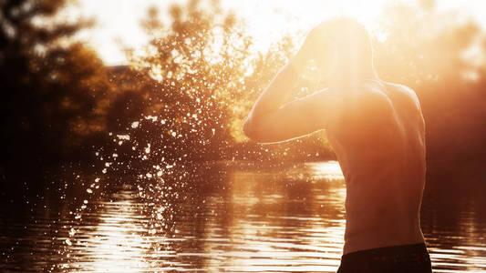 一个年轻人站在河边, 在日落时洗脸。一个裸露躯干的在水里