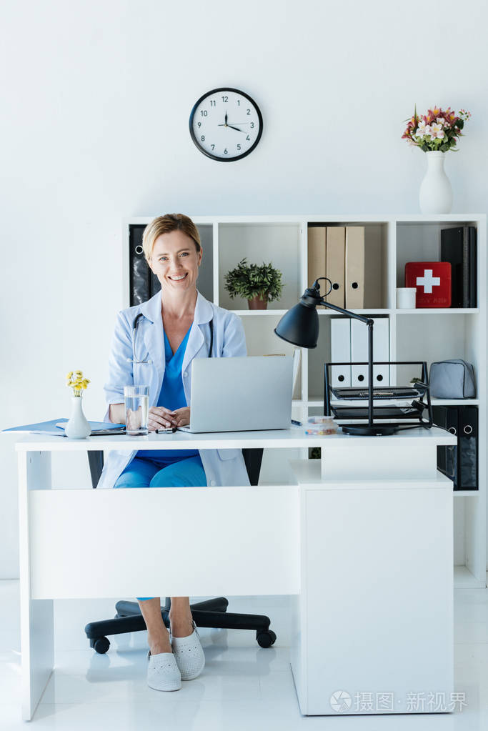 有吸引力的成年女医生看着相机和坐在桌子上的笔记本电脑在办公室