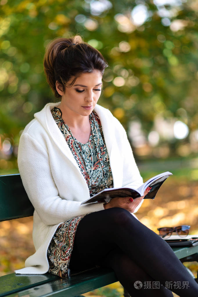 在一个阳光明媚的秋日, 美丽的年轻女子坐在公园的长凳上看书