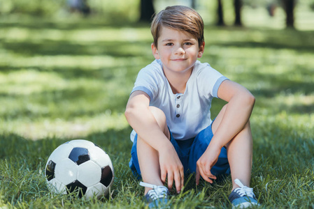 可爱的小男孩坐在草地上与足球和微笑的相机在公园