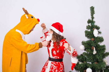 假期, 圣诞节和笑话概念滑稽的男子在鹿服装和圣诞老人服装的妇女在圣诞树上白色背景