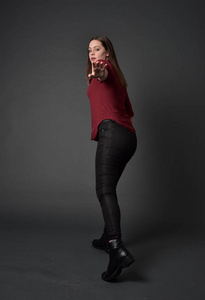穿红色衬衫和皮裤的黑发女孩的全长肖像。站在姿势, 在灰色的工作室背景