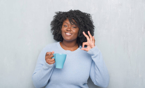 年轻的非洲裔美国妇女灰色垃圾墙喝一杯咖啡做 ok 标志与手指, 优秀的标志