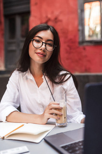 美丽的女孩与深色的头发在白色衬衫和眼镜朦胧地看着在相机与笔记本电脑和记事本在舒适的庭院咖啡馆