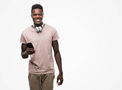 年轻的非洲裔美国人戴着耳机, 手持智能手机站着, 微笑着微笑着露出牙齿