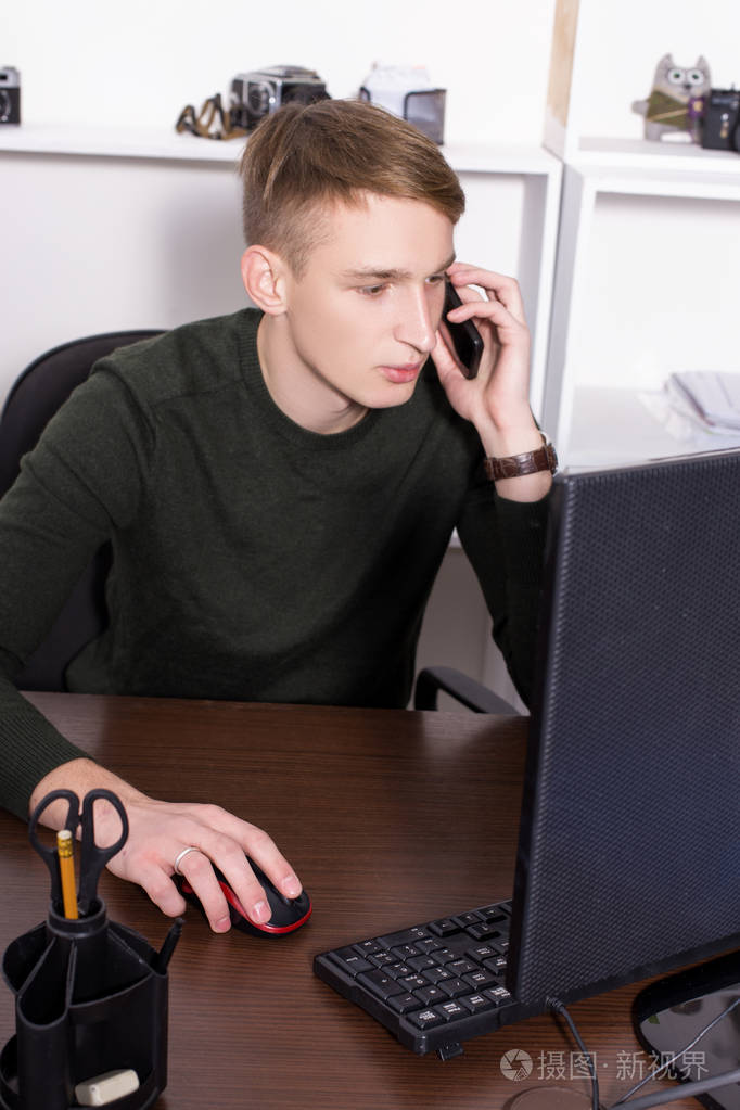 一个穿着深色毛衣的年轻人在办公室里用电脑和电话交谈.经营理念
