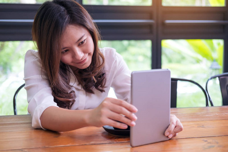 一个美丽的亚洲妇女与笑脸手持平板电脑在绿色自然背景下的现代咖啡馆中的应用
