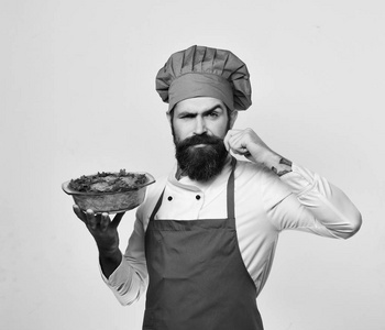 厨师与严肃的面孔在勃艮第制服举行烘烤的菜。人与胡子被隔绝在白色背景