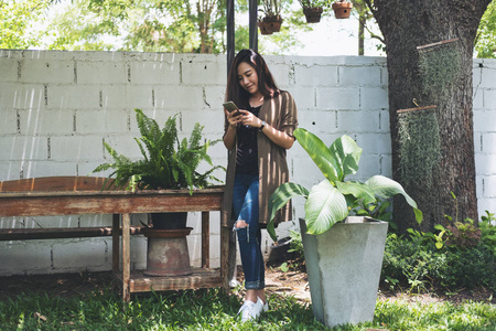 特写肖像图片一个美丽的亚洲妇女站在和使用智能手机与绿色自然和白色砖墙背景