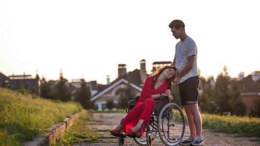 女孩在轮椅上可爱的与她的年轻人交谈在房子的背景, 绿草和树木