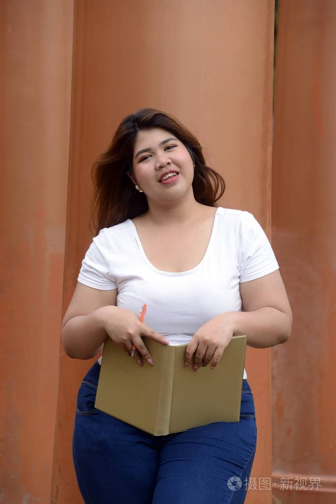 亚洲漂亮笑脸脸胖女人的肖像站在公园里的柱子上拿着一本小册子