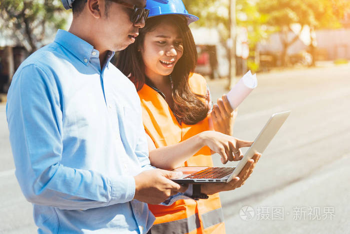 亚洲男女工程师与蓝色安全头盔会议在建筑工地与蓝图和笔记本电脑