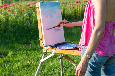 一位年轻的女艺术家拿着画笔, 在日落的光线下在画架上画一幅画。画家在日落时在花园里画油画, 特写