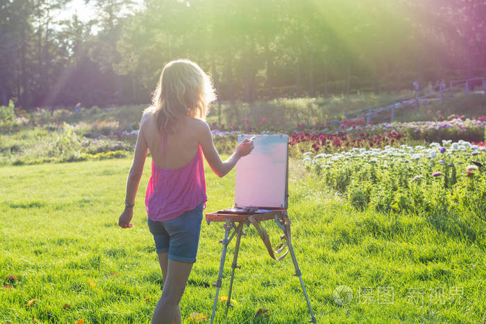 一位年轻的女艺术家拿着画笔, 在日落的光线下在画架上画一幅画。画家在日落时在花园里画油画。
