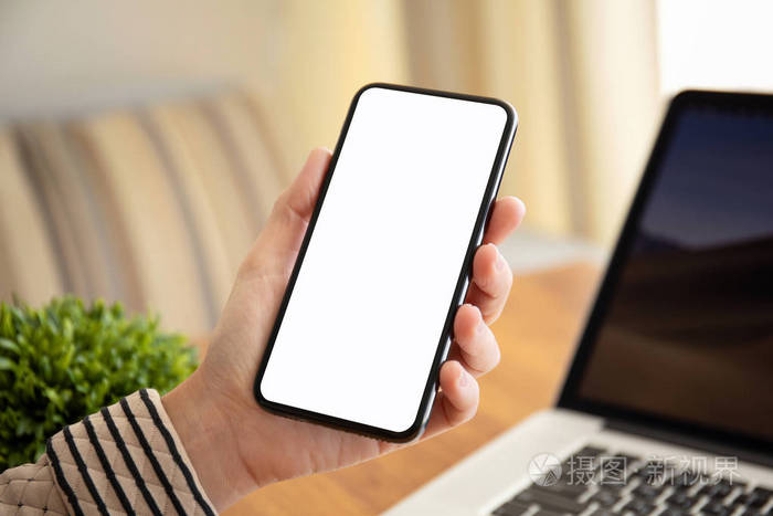 女性手拿着触摸手机与孤立的屏幕以上的桌子与笔记本电脑在办公室