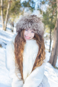 女孩穿着白色毛皮大衣和毛皮帽子, 蓝眼睛和长头发的背景下的雪和树木。冬天