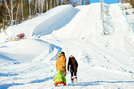 在阳光明媚的冬日午后, 一对夫妇朝雪坡走去, 和他们的小儿子一起拉雪橇。