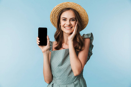 一个满意的年轻女子在礼服和夏季帽子显示在蓝色背景下隔离的空白屏幕手机的肖像
