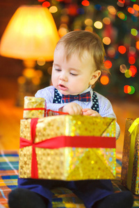 有趣的婴儿坐在地板上, 手里拿着一份大礼物, 背景是明亮的节日灯光