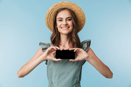 一个开朗的年轻女子在礼服和夏季帽子显示空白屏幕手机在蓝色背景下隔离的肖像