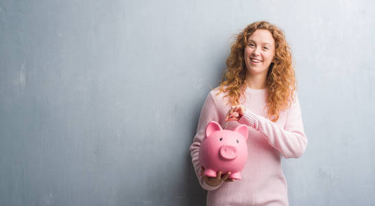 灰色垃圾墙上的年轻红头发的女人持有小猪银行和美元的快乐的脸站在微笑着自信的微笑显示牙齿