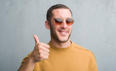 年轻的白种人在灰色的垃圾墙上戴着太阳镜快乐的大微笑做 ok 标志, 拇指与手指, 优秀的标志