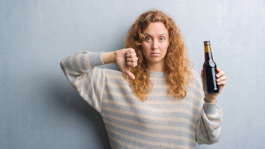 灰色垃圾墙上的年轻红头发的女人拿着啤酒瓶与愤怒的脸, 消极的迹象显示不喜欢拇指下来, 拒绝概念