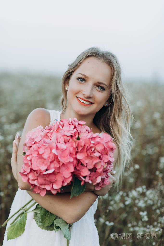 美丽的金发女孩, 蓝色的眼睛和长发, 捧着一束粉红色的花朵和微笑